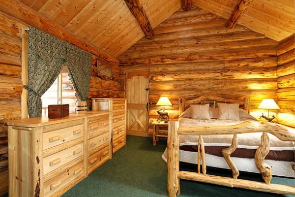 Warm cozy bedroom with rustic bed, nightstand and dresser. Green carpet floor and curtains. Log cabin house interior